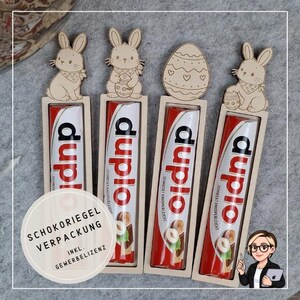 Op de afbeelding: Houten paas-chocoladereep-houders met Duplo-repen. Elke houder heeft een lasergesneden konijn- of ei-ontwerp. De chocoladerepen zijn rood en wit met het Duplo-logo. De tekst "SCHOKORIEGEL VERPACKUNG" is inbegrepen.
