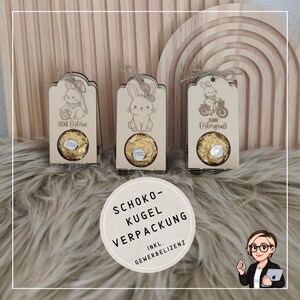 Op de afbeelding: Drie houten paasgeschenkdozen met lasergesneden konijnenontwerpen, elk met een gouden chocoladesfeer. De dozen hebben verschillende konijnenillustraties en tekst. Een ronde sticker met Duitse tekst staat op de voorgrond.