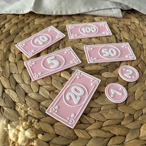 May include: Pink play money bills and coins with white borders and numbers. The bills are in denominations of 5, 10, 20, 50, and 100. The coins are marked with 1 and 2. Each bill and coin has a heart design.