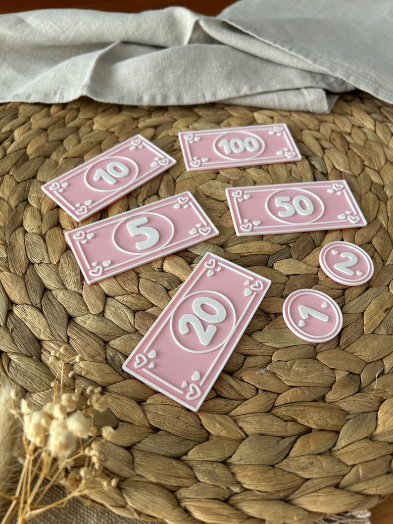 May include: Pink play money bills and coins with white borders and numbers. The bills are in denominations of 5, 10, 20, 50, and 100. The coins are marked with 1 and 2. Each bill and coin has a heart design.