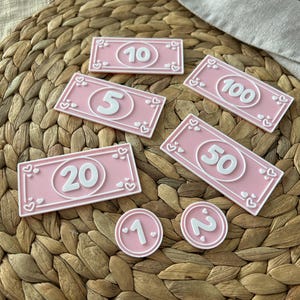 May include: Pink play money in various denominations, including $1, $2, $5, $10, $20, $50, and $100 bills. Each bill and coin features a white number and heart-shaped accents. The items are arranged on a woven surface.