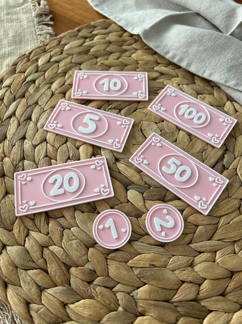 May include: Pink play money in various denominations, including $1, $2, $5, $10, $20, $50, and $100 bills. Each bill and coin features a white number and heart-shaped accents. The items are arranged on a woven surface.