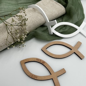 May include: A table setting with a linen napkin, white fish-shaped napkin rings, and wooden fish-shaped place cards. The place cards are engraved with the name "Magdalena" and the date "21.08.2024". The background includes green fabric and small white flowers.