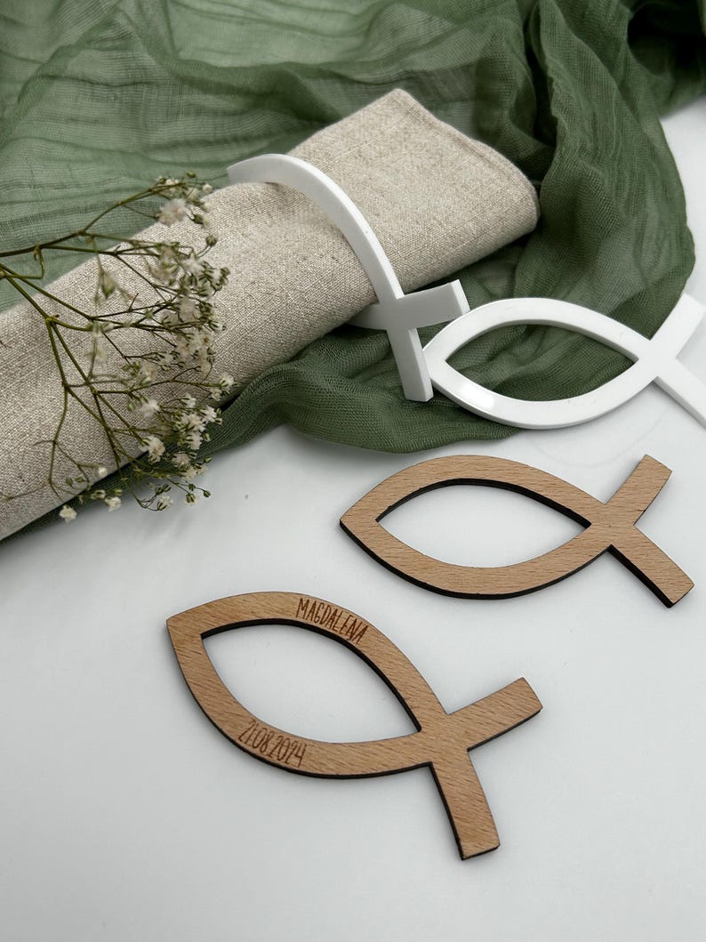 May include: A table setting with a linen napkin, white fish-shaped napkin rings, and wooden fish-shaped place cards. The place cards are engraved with the name "Magdalena" and the date "21.08.2024". The background includes green fabric and small white flowers.