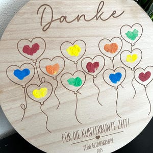 Puede incluir: Una placa redonda de madera con la palabra "Danke" en cursiva. Presenta globos en forma de coraz&oacute;n en varios colores, incluyendo rojo, amarillo y azul. Tambi&eacute;n est&aacute; presente el texto "F&Uuml;R DIE KUNTERBUNTE ZEIT!" y "DEINE BLUMENGRUPPE 2025".