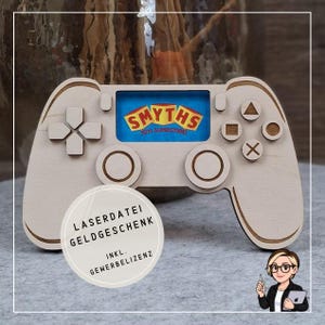 Puede incluir: Un mando de videojuegos de madera con una pantalla azul que muestra el logotipo "SMYTHS TOYS SUPERSTORES". El mando tiene botones y una cruceta. Una pegatina redonda con texto en alemán está en la esquina inferior izquierda.