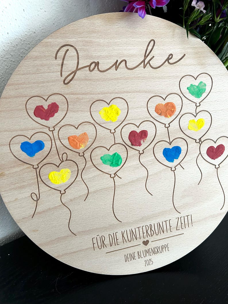 Puede incluir: Una placa redonda de madera con la palabra "Danke" en cursiva. Presenta globos en forma de coraz&oacute;n en varios colores, incluyendo rojo, amarillo y azul. Tambi&eacute;n est&aacute; presente el texto "F&Uuml;R DIE KUNTERBUNTE ZEIT!" y "DEINE BLUMENGRUPPE 2025".