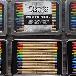 May include: Three sets of 12 watercolour pencils in metal tins. Each tin has a black and white label that reads "Distress Watercolour Pencils 12 Woodless Water-Reactive Highly Pigmented Pencils".