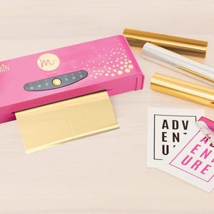 Puede incluir: Máquina de prensa de calor Pink-Miah con una lámina de lámina dorada y una hoja de papel con la palabra "Aventura" impresa en ella.