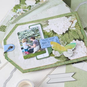 Puede incluir: Una etiqueta de álbum de recortes hecha a mano con una foto de una persona y un niño, adornada con una mariposa y detalles florales. La etiqueta presenta un cierre azul y troqueles de metal decorativos. La etiqueta es verde y blanca.