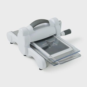 May include: A white die-cutting and embossing machine with a gray handle and a star-shaped die. The machine has a gray roller handle and a clear plastic platform. The text "Big Shot" is visible on the side.