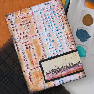 May include: A textured art piece with a rectangular shape, featuring a mix of orange, red, and blue hues. The design includes a small black rectangle with the word "Storyteller" printed on it. A roller and paint palette are visible.