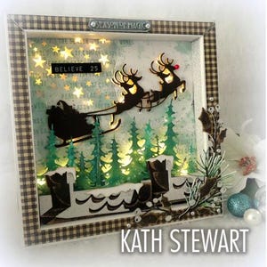 May include: A framed Christmas shadow box featuring a silhouette of Santa's sleigh and reindeer over a forest of green trees. The box has a plaid border and the words "SEASON OF MAGIC" and "BELIEVE 25". The background has star-shaped lights.