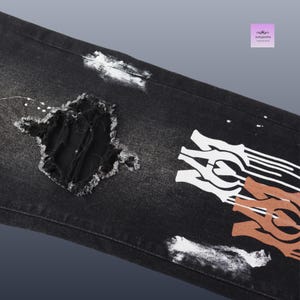 May include: Black denim jeans with distressed details, including a large ripped section and white paint splatters. The jeans feature a white and brown graphic design on the leg.