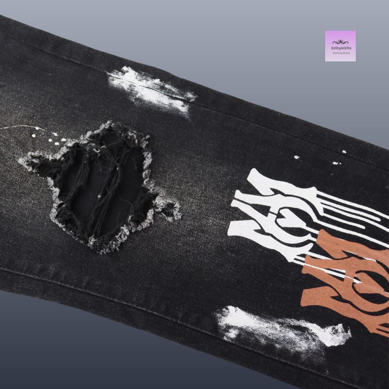 May include: Black denim jeans with distressed details, including a large ripped section and white paint splatters. The jeans feature a white and brown graphic design on the leg.