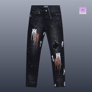 May include: Black distressed jeans with white and rust-colored paint splatter and logo details. The jeans have a ripped knee and a button closure. A tag is attached to the waistband.