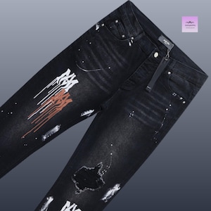 May include: Black denim jeans with white and rust-colored paint splatter and distressed details. The jeans feature a button closure, a zip fly, and the brand name "AMIRI" on the tag.