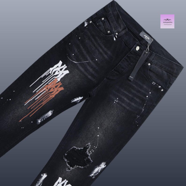 May include: Black denim jeans with white and rust-colored paint splatter and distressed details. The jeans feature a button closure, a zip fly, and the brand name "AMIRI" on the tag.
