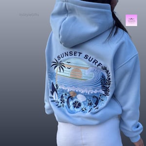 May include: Pale blue hoodie with a "Sunset Surf" design on the back. The graphic includes a palm tree, waves, and the words "Sunset Surf". The hoodie has a hood and long sleeves. The brand name "ItsStyleGifts" is visible.