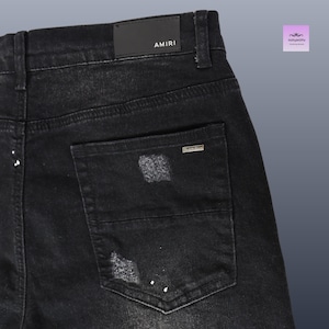 May include: Close-up of black denim jeans with distressed details and white paint splatters. The back pocket features a small metal tag and the brand name "AMIRI" is visible on the waistband.