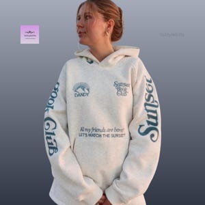 May include: A light grey hoodie featuring teal text that reads "Sunset Book Club" and "Dandy". The sleeves display "Sunset" and "Book Club". The hoodie also has the text "All my friends are here! Let's watch the sunset."