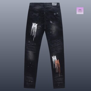 May include: Black denim jeans with distressed details and white and brown paint splatter accents. The jeans feature a unique design with a logo on the legs. A stylish and edgy fashion item.