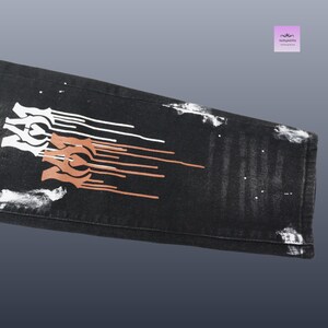 May include: Black denim jeans with white and brown abstract designs. The jeans have a distressed look with white paint splatters. The design is on the lower leg of the jeans.