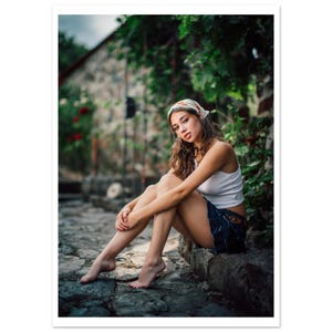 May include: A woman wearing a white tank top, denim shorts, and a patterned headscarf sits on a stone surface. The background features a stone building and lush greenery. The image has a soft, natural lighting.