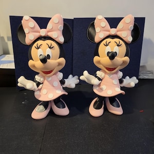 Pu&ograve; includere: Due statuette di Minnie Mouse. Ogni statuetta ha una testa nera con grandi orecchie, un fiocco rosa a pois bianchi e un vestito rosa a pois bianchi. Le statuette sono in piedi con le braccia tese e indossano scarpe rosa.