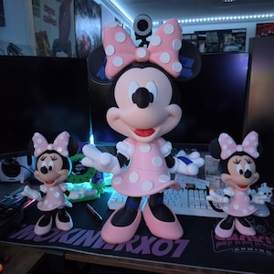 Pu&ograve; includere: Tre statuette di Minnie Mouse di diverse dimensioni. Le figure indossano abiti rosa con pois bianchi e fiocchi abbinati. La figura pi&ugrave; grande &egrave; al centro, affiancata da due versioni pi&ugrave; piccole. Le figure sono su una scrivania con attrezzature informatiche.