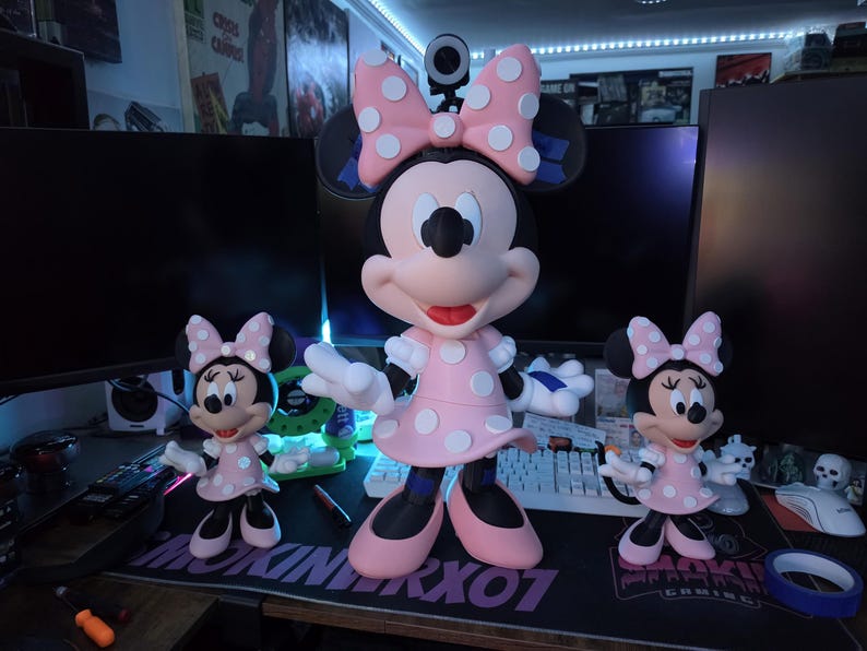 Pu&ograve; includere: Tre statuette di Minnie Mouse di diverse dimensioni. Le figure indossano abiti rosa con pois bianchi e fiocchi abbinati. La figura pi&ugrave; grande &egrave; al centro, affiancata da due versioni pi&ugrave; piccole. Le figure sono su una scrivania con attrezzature informatiche.