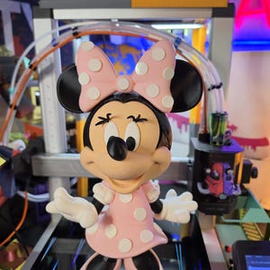 Pu&ograve; includere: Una statuetta di Minnie Mouse, con un vestito rosa a pois bianchi e un fiocco abbinato. La figura ha orecchie nere, un naso nero e guanti bianchi. La statuetta &egrave; su una superficie nera.