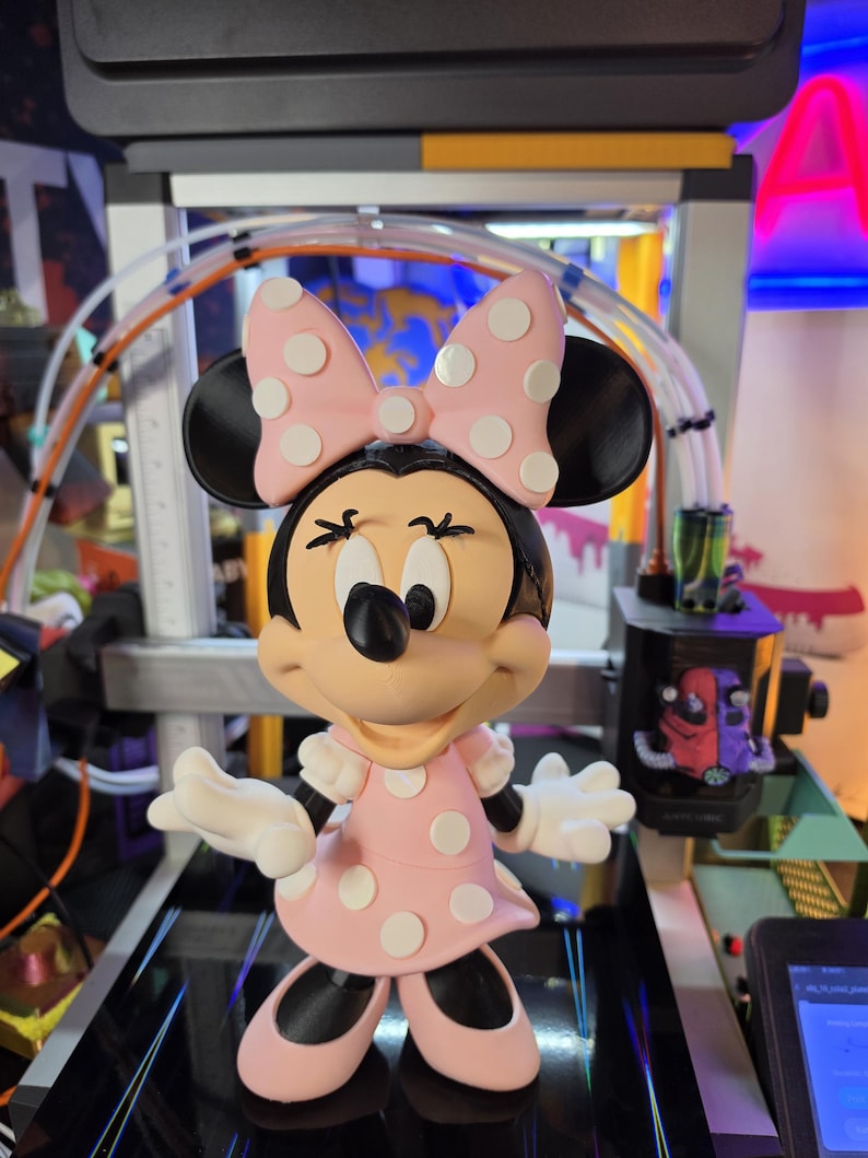 Pu&ograve; includere: Una statuetta di Minnie Mouse, con un vestito rosa a pois bianchi e un fiocco abbinato. La figura ha orecchie nere, un naso nero e guanti bianchi. La statuetta &egrave; su una superficie nera.