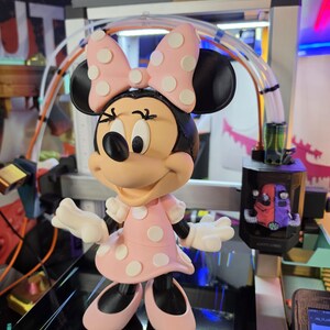 Pu&ograve; includere: Una statuetta di Minnie Mouse, con un vestito e un fiocco rosa a pois bianchi. La figura ha orecchie, scarpe e un viso sorridente neri. La statuetta &egrave; su una superficie nera.