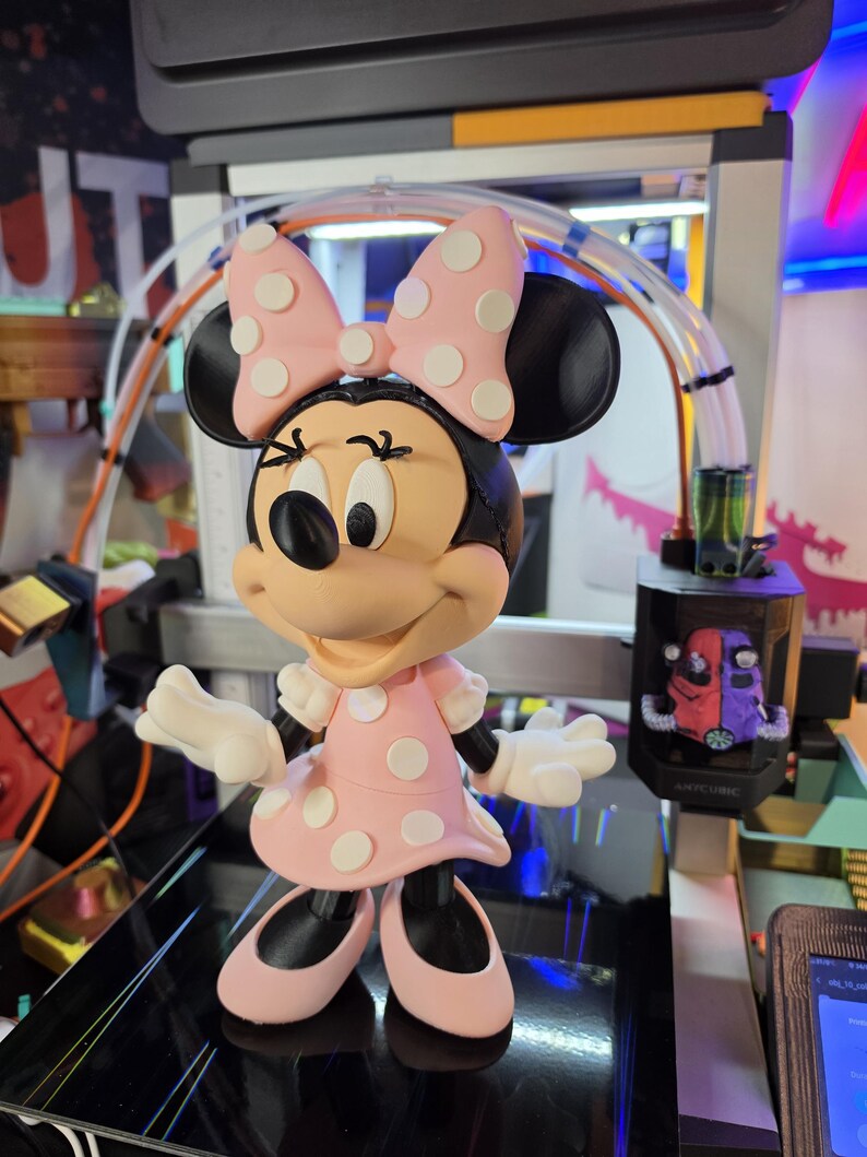 Pu&ograve; includere: Una statuetta di Minnie Mouse, con un vestito e un fiocco rosa a pois bianchi. La figura ha orecchie, scarpe e un viso sorridente neri. La statuetta &egrave; su una superficie nera.