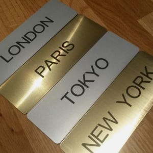 Puede incluir: Cuatro letreros rectangulares de metal con los nombres de ciudades: Londres, París, Tokio y Nueva York. Los letreros de Londres y Tokio son plateados, mientras que los de París y Nueva York son dorados. El texto es negro.