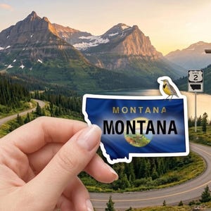 May include: A blue and white Montana state sticker with a bird perched on top. The sticker is held in front of a scenic landscape of mountains, a lake, and a road. The sticker has the word "MONTANA" on it.