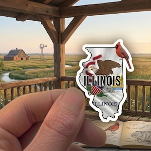 Puede incluir: Una mano sosteniendo una pegatina de la bandera de Illinois con un cardenal. El fondo presenta una estructura de madera, un letrero que dice "Prairie State Overlook" y un paisaje rural con un granero y un molino de viento.