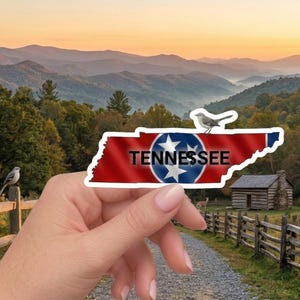 May include: A sticker in the shape of Tennessee's outline, featuring the state flag design with the word "TENNESSEE". The sticker is held in front of a scenic landscape with mountains, trees, and a sign that reads "TENNESSEE: THE VOLUNTEER STATE - EST. 1796".
