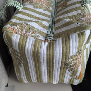 May include: Quilted tote bag with a striped pattern in white and olive green. The bag features a repeating design of stylized trees in orange and green. The handles and zipper trim are green.