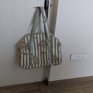May include: A tote bag with vertical stripes in white and olive green, adorned with palm tree motifs. The bag has matching striped handles and is hanging on a black hook. The bag is made of a soft fabric.