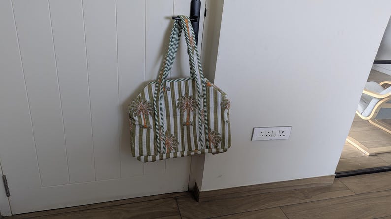 May include: A tote bag with vertical stripes in white and olive green, adorned with palm tree motifs. The bag has matching striped handles and is hanging on a black hook. The bag is made of a soft fabric.