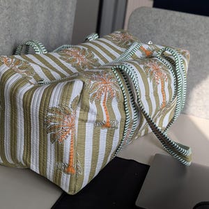 May include: A rectangular tote bag with a striped pattern in white and olive green, featuring palm tree illustrations in orange and brown. The bag has matching handles and a striped strap, perfect for carrying essentials.