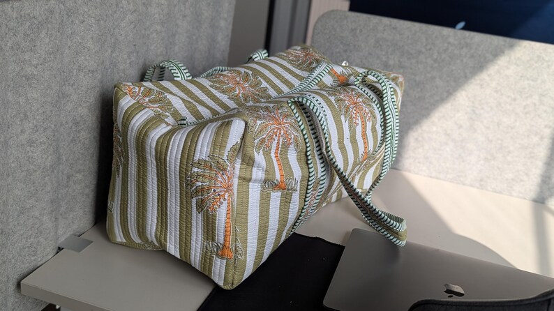 May include: A rectangular tote bag with a striped pattern in white and olive green, featuring palm tree illustrations in orange and brown. The bag has matching handles and a striped strap, perfect for carrying essentials.
