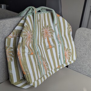 May include: A tote bag with a striped pattern in white, olive green, and orange. The bag features a repeating palm tree design and green trim. The handles are made of a twisted rope-like material.