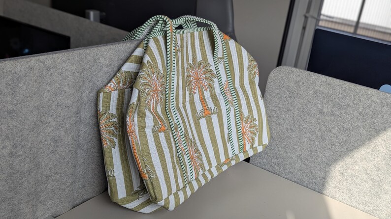 May include: A tote bag with a striped pattern in white, olive green, and orange. The bag features a repeating palm tree design and green trim. The handles are made of a twisted rope-like material.