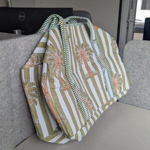 May include: A quilted tote bag with a striped pattern in white and olive green, featuring palm tree motifs in orange. The bag has a green trim and handles, and is folded over.