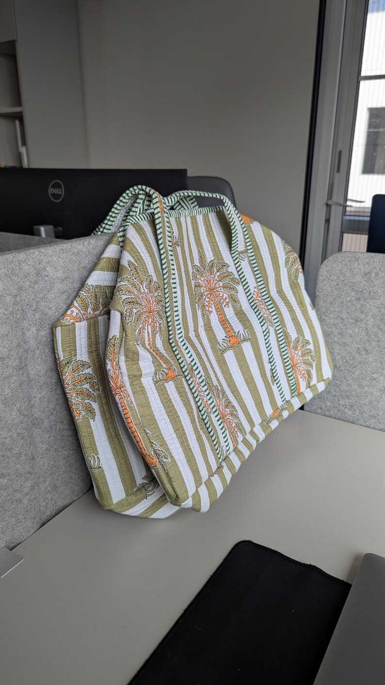May include: A quilted tote bag with a striped pattern in white and olive green, featuring palm tree motifs in orange. The bag has a green trim and handles, and is folded over.