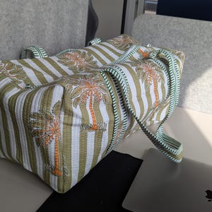 May include: A large tote bag with a striped pattern in white and olive green, featuring palm tree illustrations. The bag has matching handles and trim in a light green color. The bag is sitting on a white surface.