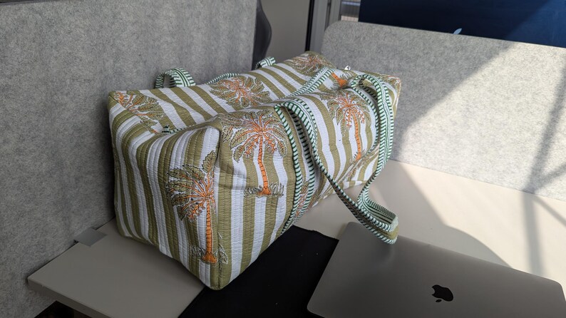 May include: A large tote bag with a striped pattern in white and olive green, featuring palm tree illustrations. The bag has matching handles and trim in a light green color. The bag is sitting on a white surface.